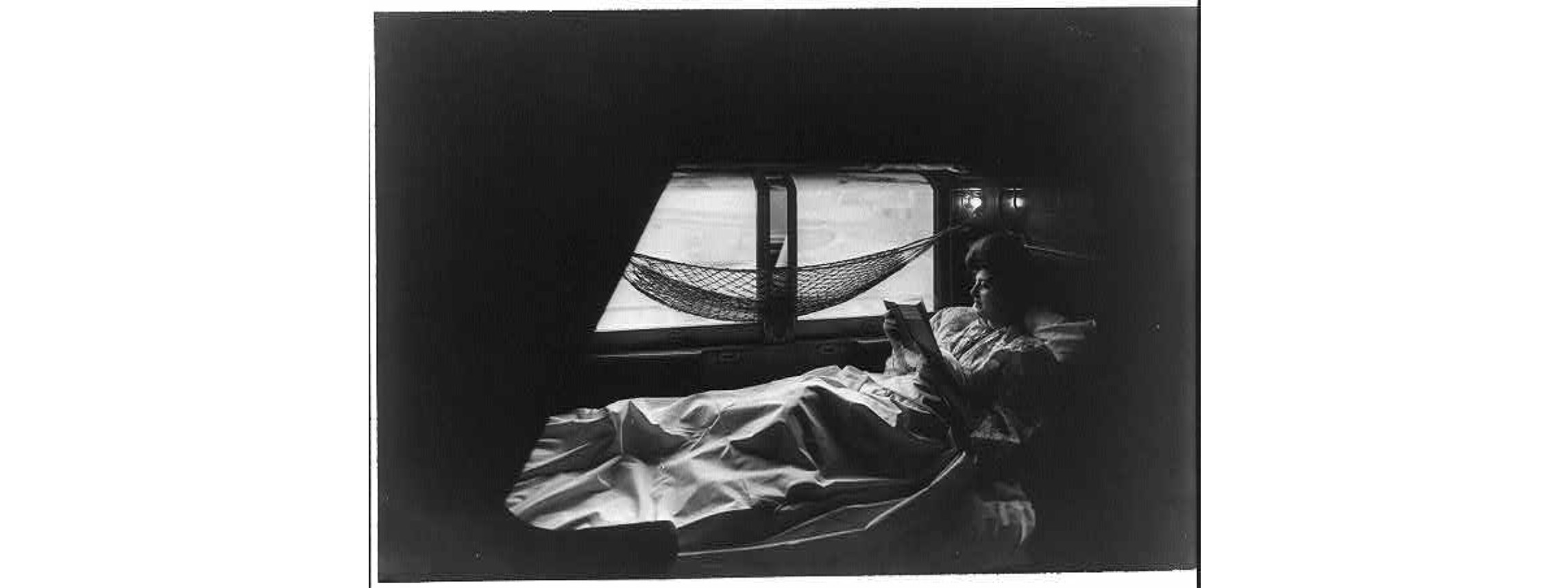 Black and white photo of a woman in early 20th century clothing reading while lying down in a sleeping birth on a train. A small hammock for belongings hands abvoe her.