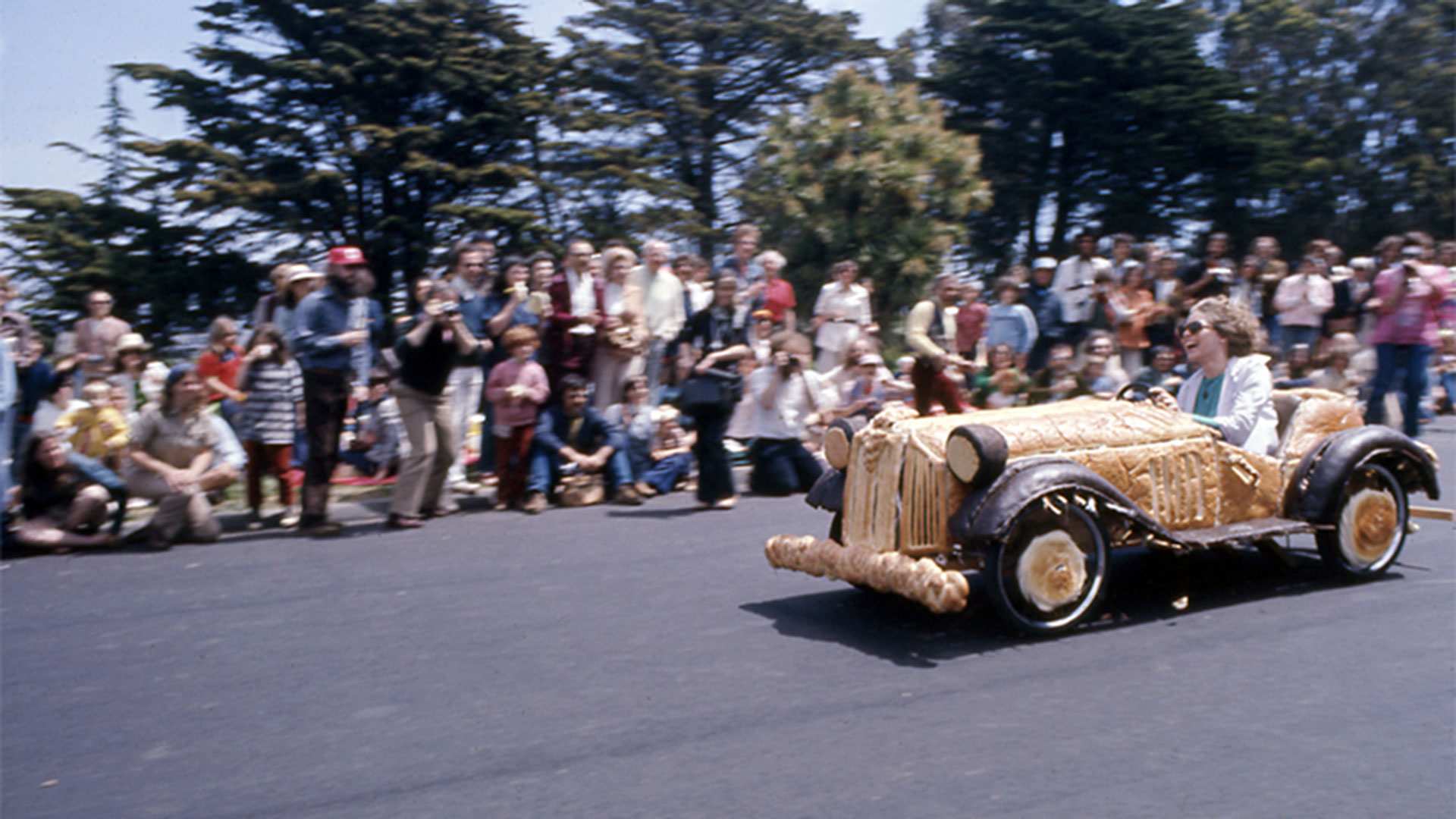 A woman in a car entirely made out of bread comes speeding down a hill. A crowd of spectators look on with trees behind them.