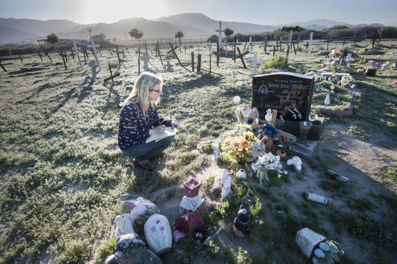 A women kneels down in a cemetery, and writes on a pad.