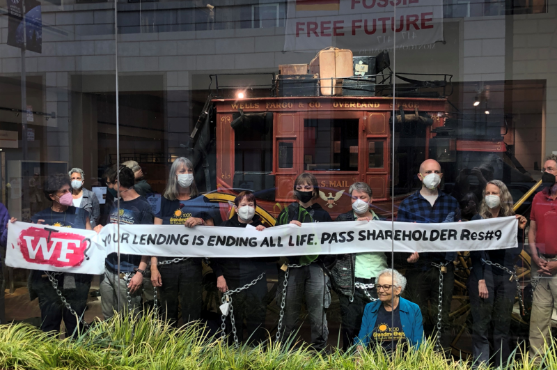 A group of protestors stands shoulder to shoulder on the far side of a floor-to-ceiling window, facing out to a street, including passing cars, seen reflected in the glass. Behind them is a red, antique horse carriage with "Wells Fargo & Co. Overland Stage" lettered in yellow across the top, and luggage piled on the roof. Beyond the carriage is a sign hanging from a second-story walkway that says "Fossil Free Future." Nine of the visible protestors in the foreground hold a thin white banner at chest height that reads "W(t)F(?): Your lending is ending all life. Pass shareholder Res #9." All wear facemasks and appear calm, and most appear to be smiling. At first glance, most appear to be white and middle-aged.