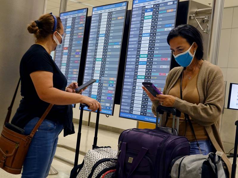Two travelers with face masks stand by a schedule board in an airport.