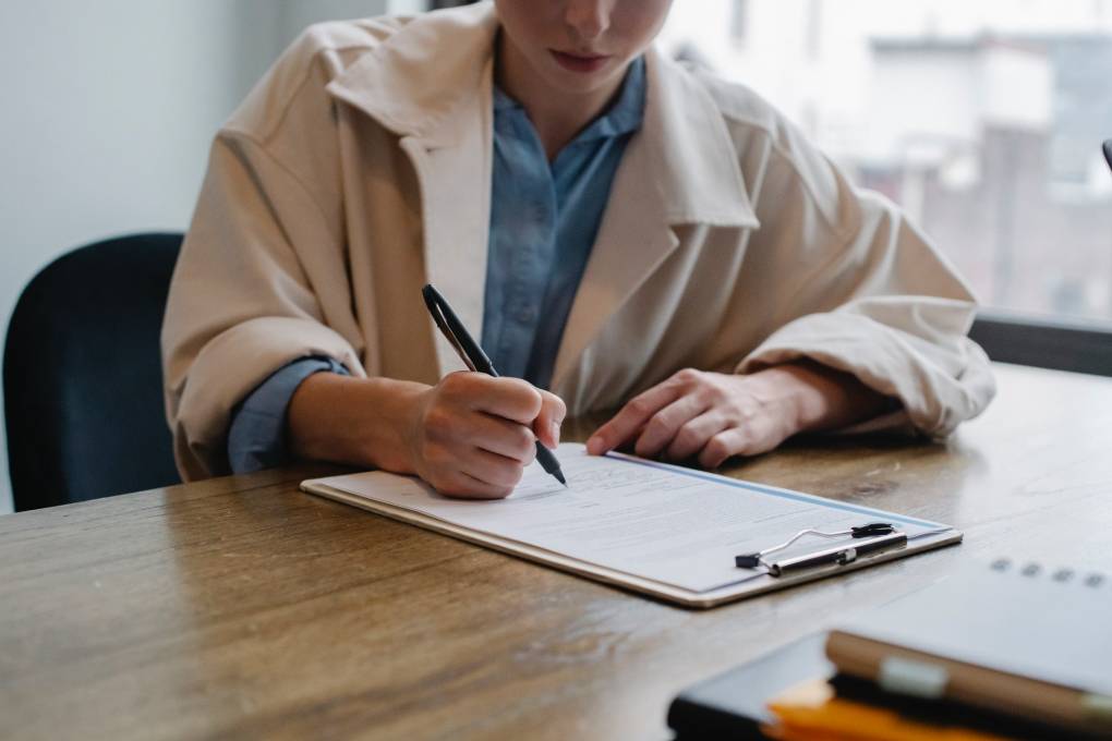 A woman in a large beige trench coat sits at a table writing on a clipboard.