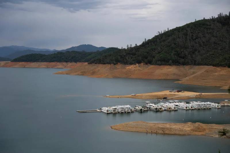 A steep embankment of brown dirt and clay borders Shasta Lake in April 2022, showing low water levels. A border of thick trees runs along the top of the brown embankment. Houseboats float at a pier.
