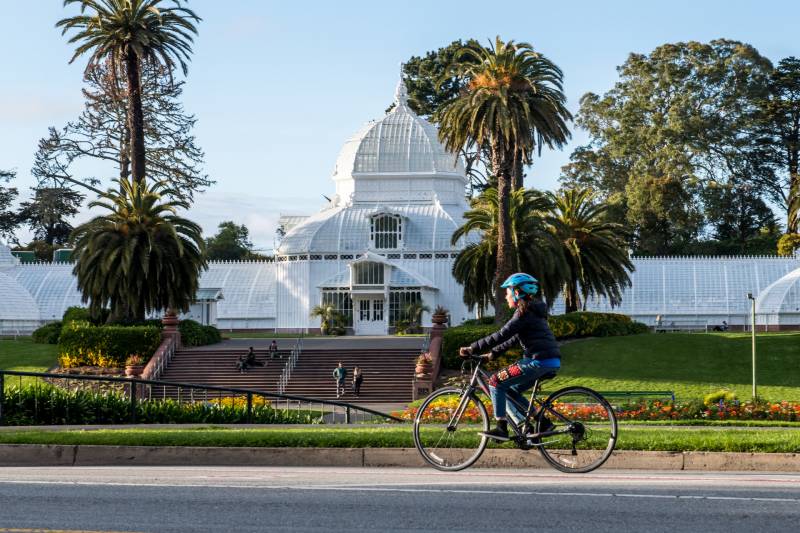 A large white building with a dome sits in the background with yellow and red flowers growing in front. A cyclist rides past in the foreground.