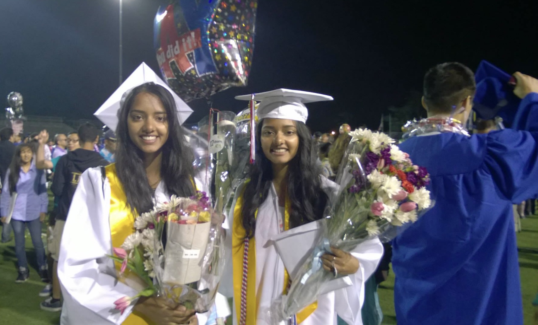 Two teenage girls dressed in graduation gowns and holding flowers smile at the camera.