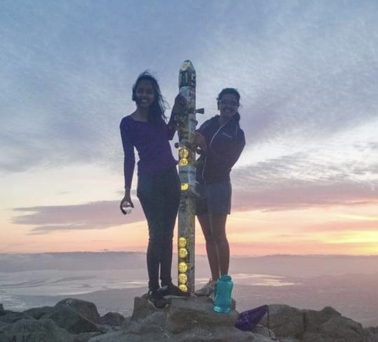 Two teenage girls stand at the top of a mountain as the sun sets behind them.