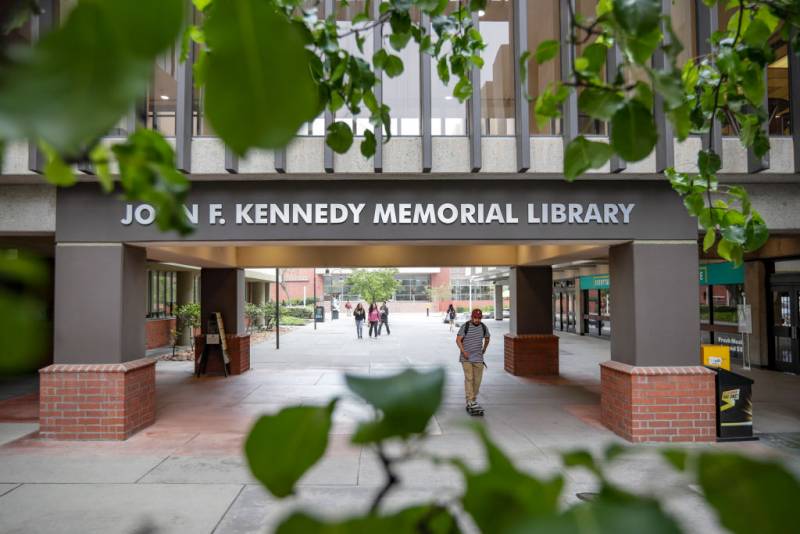 People are seen walking in the distance on a campus and under a building called "John F. Kennedy Memorial Library."