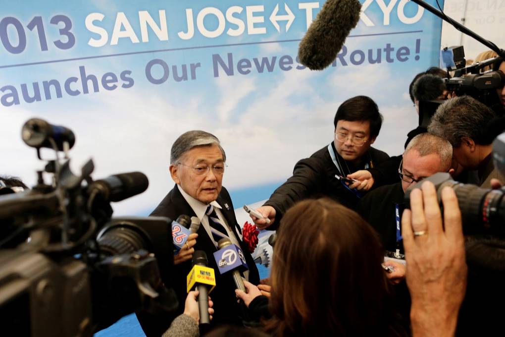 Norman Minetastands inside an airport, in front of a large sign. Many cameras and reporters surround him asking hi questions, he looks over at one member of the media.