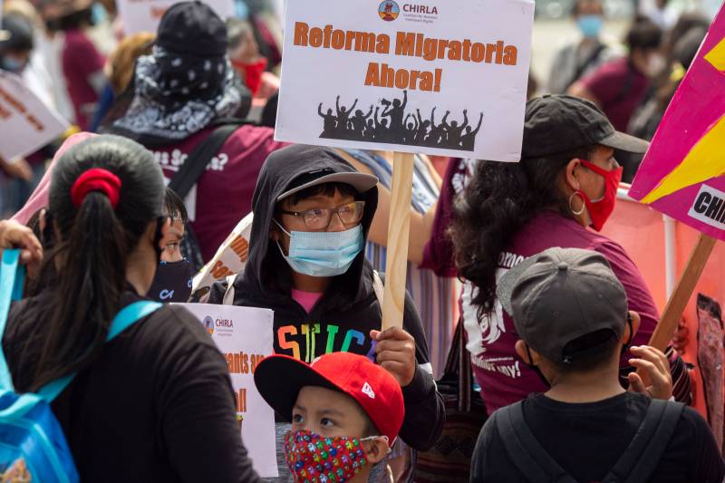 Amid a crowd of people wearing caps and face masks, one person wearing a black hoodie with the word "justice" in rainbow colors holds a sign reading "Reforma Migratoria Ahora!"