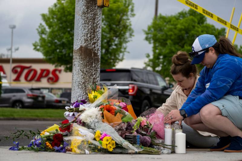 Two women kneel down to put flowers and candles on the sidewalk, in front of a Tops supermarket.