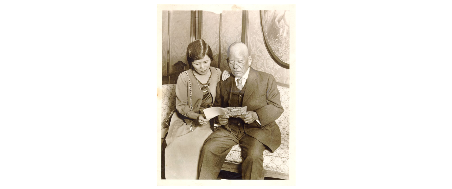 Black and white photo of an older Asian man in a suit looking at a card. A younger Asian woman reads over his shoulder. They are dressed in clothes typical of the early 20th century.
