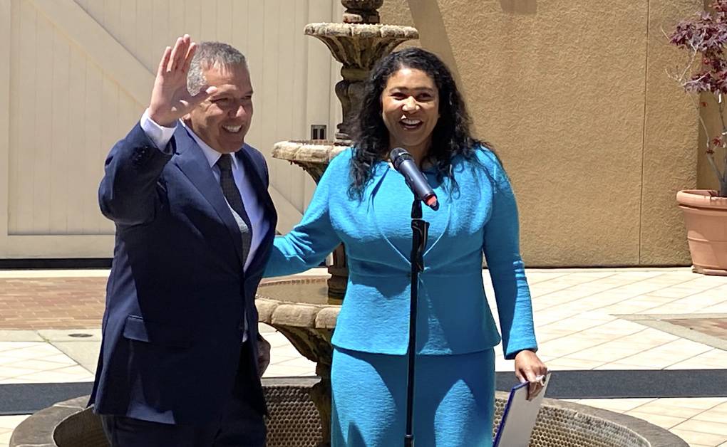 Mayor London Breed, in a blue suit, smiles with her hand on the shoulder of a man.