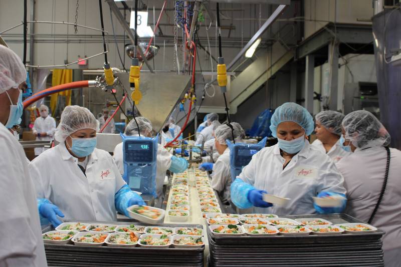 Several people wearing white oats, light blue hair nets and face masks in front of a conveyer belt of plates with food in a factory.