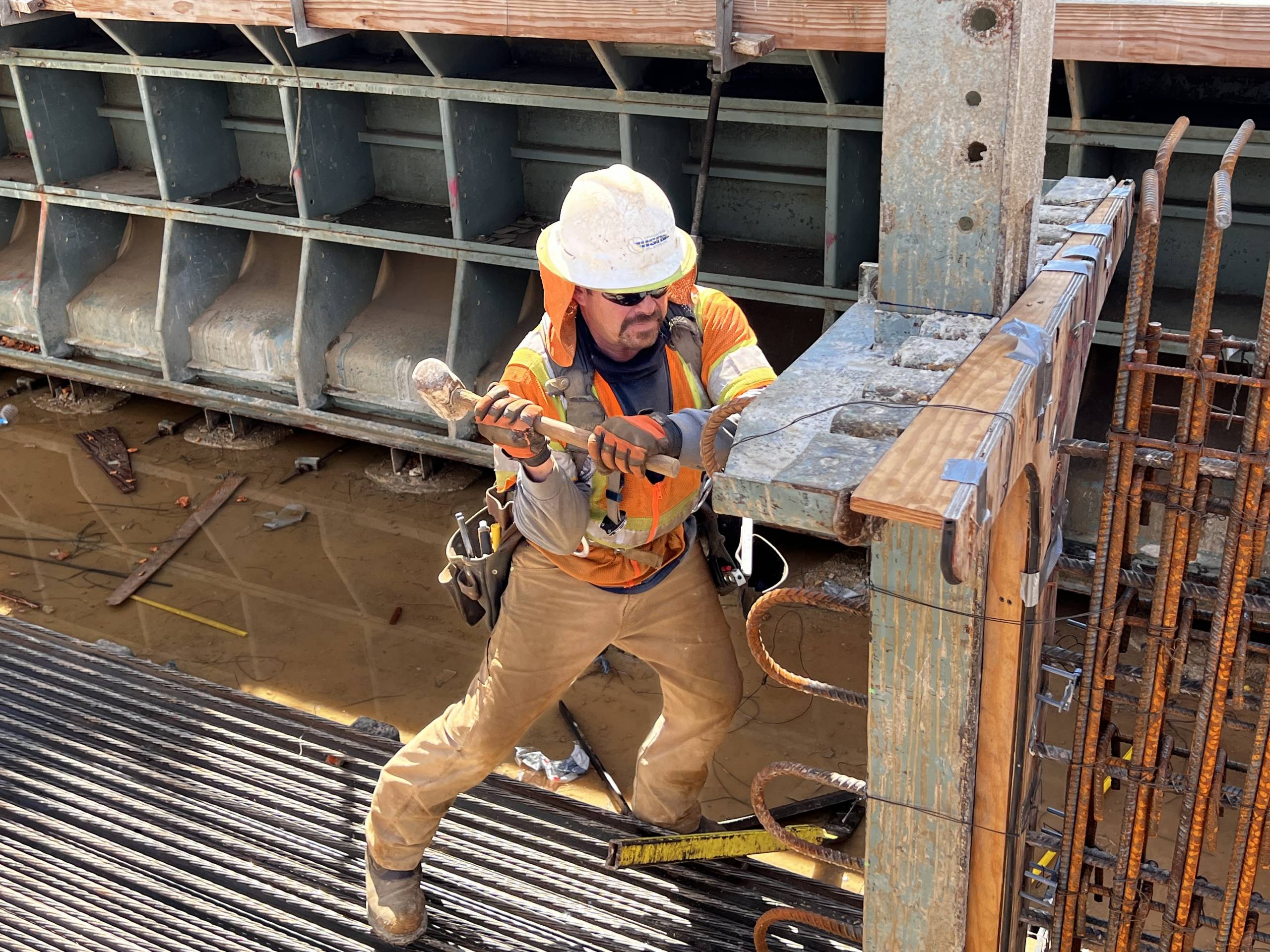 A construction work in an orange vest and white hard hat hammers a steel beam.