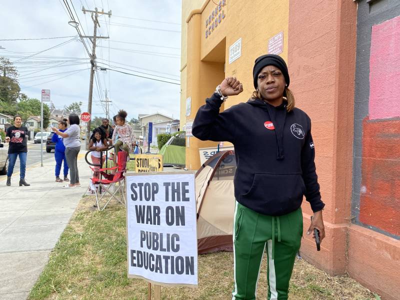 I woman stands outside a building with her fist up. A sign next to her says "stop the war on public education."