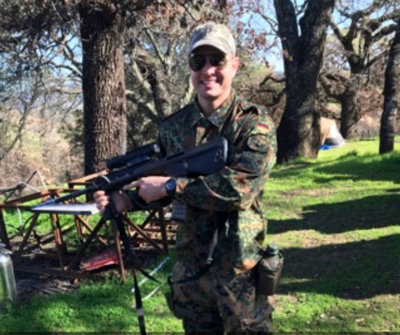 man wearing military fatigues and sunglasses outdoors smiles as he holds what appears to be an assault rifle