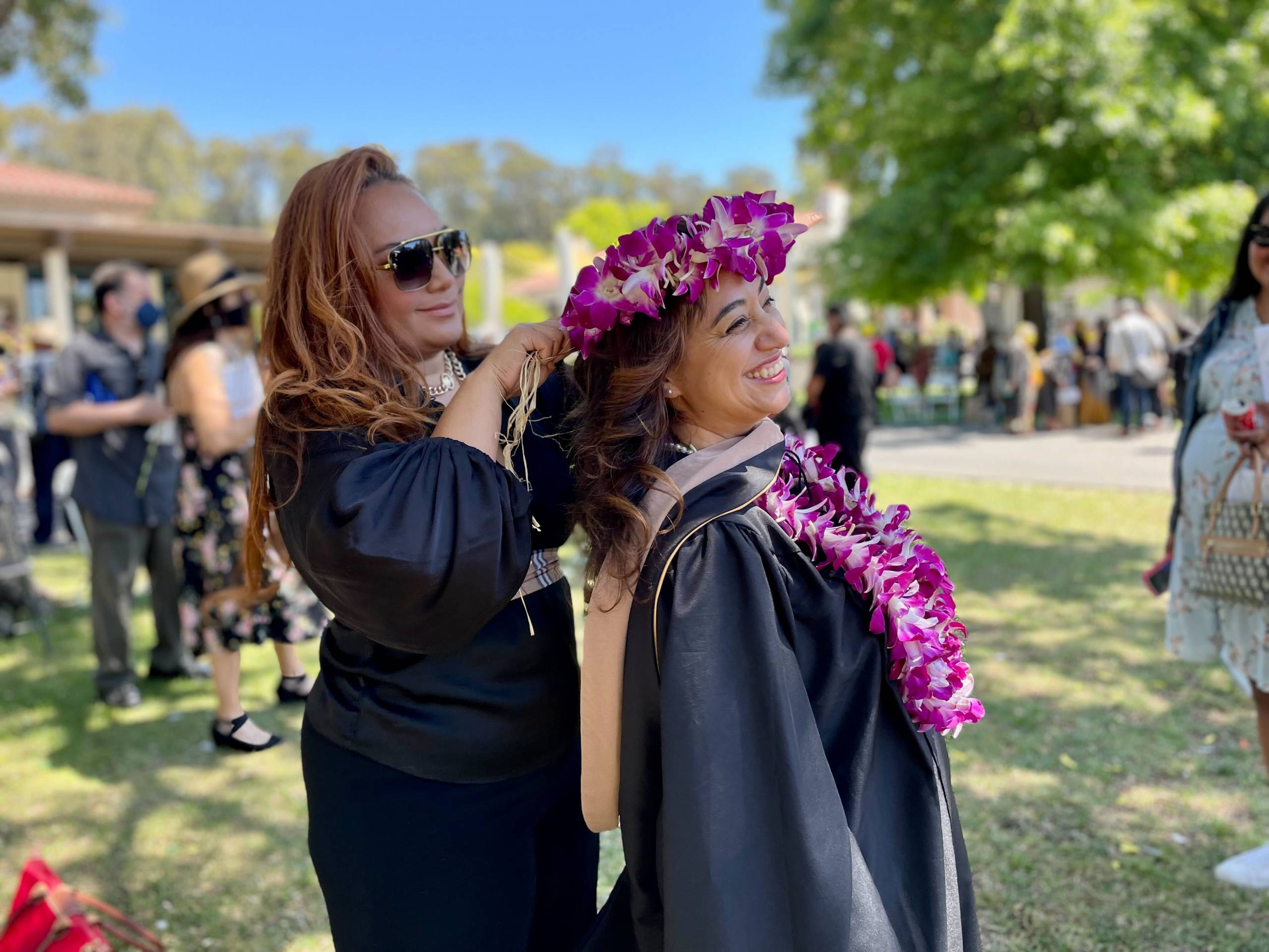 A woman ties a colorful purple lei onto another woman wearing a black graduation gown. A sunny day with clear sky and trees can be seen in the background.