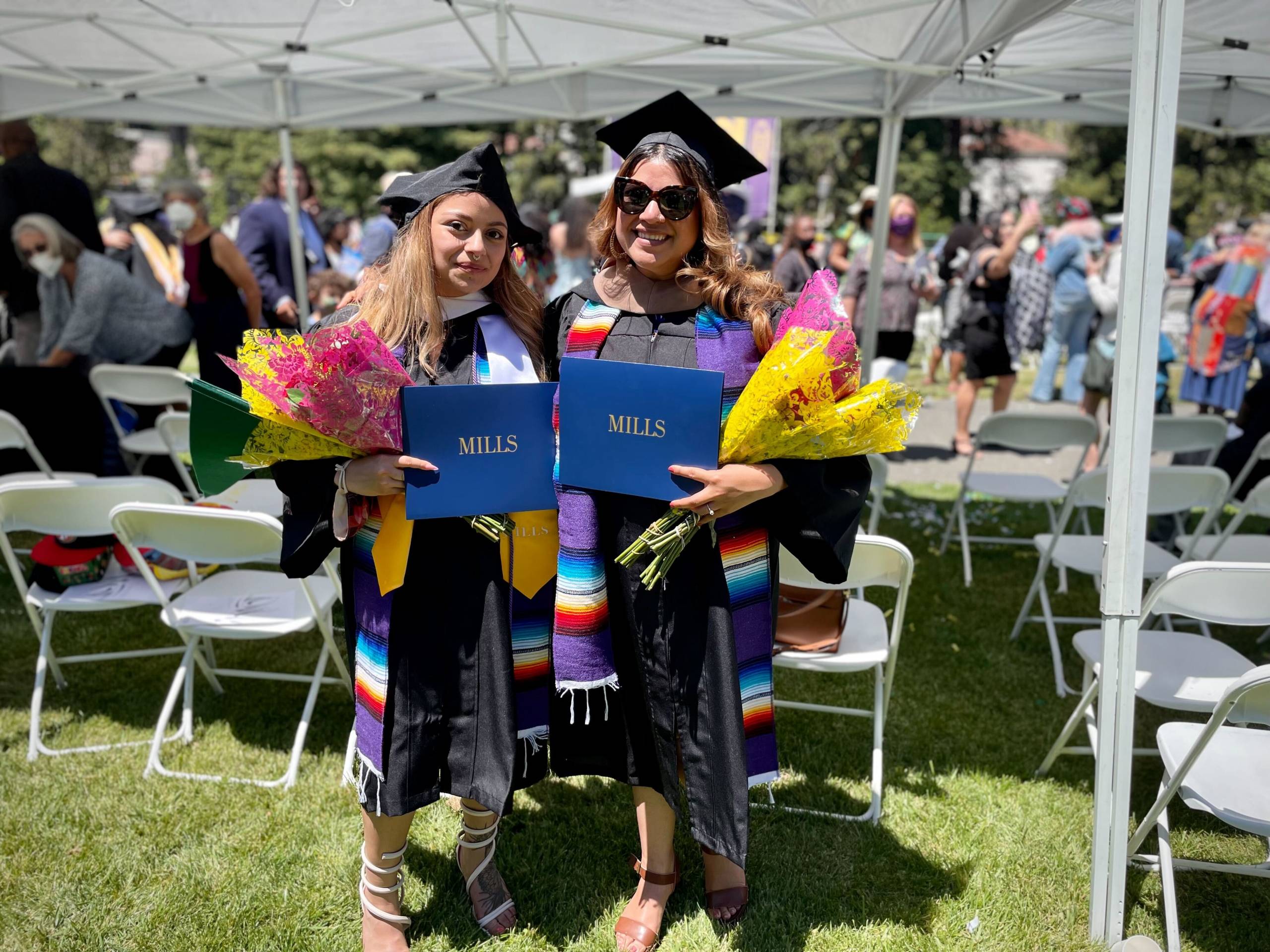 Two people holding their Mills College degrees stand in black cap and gowns holding bouquets of flowers.