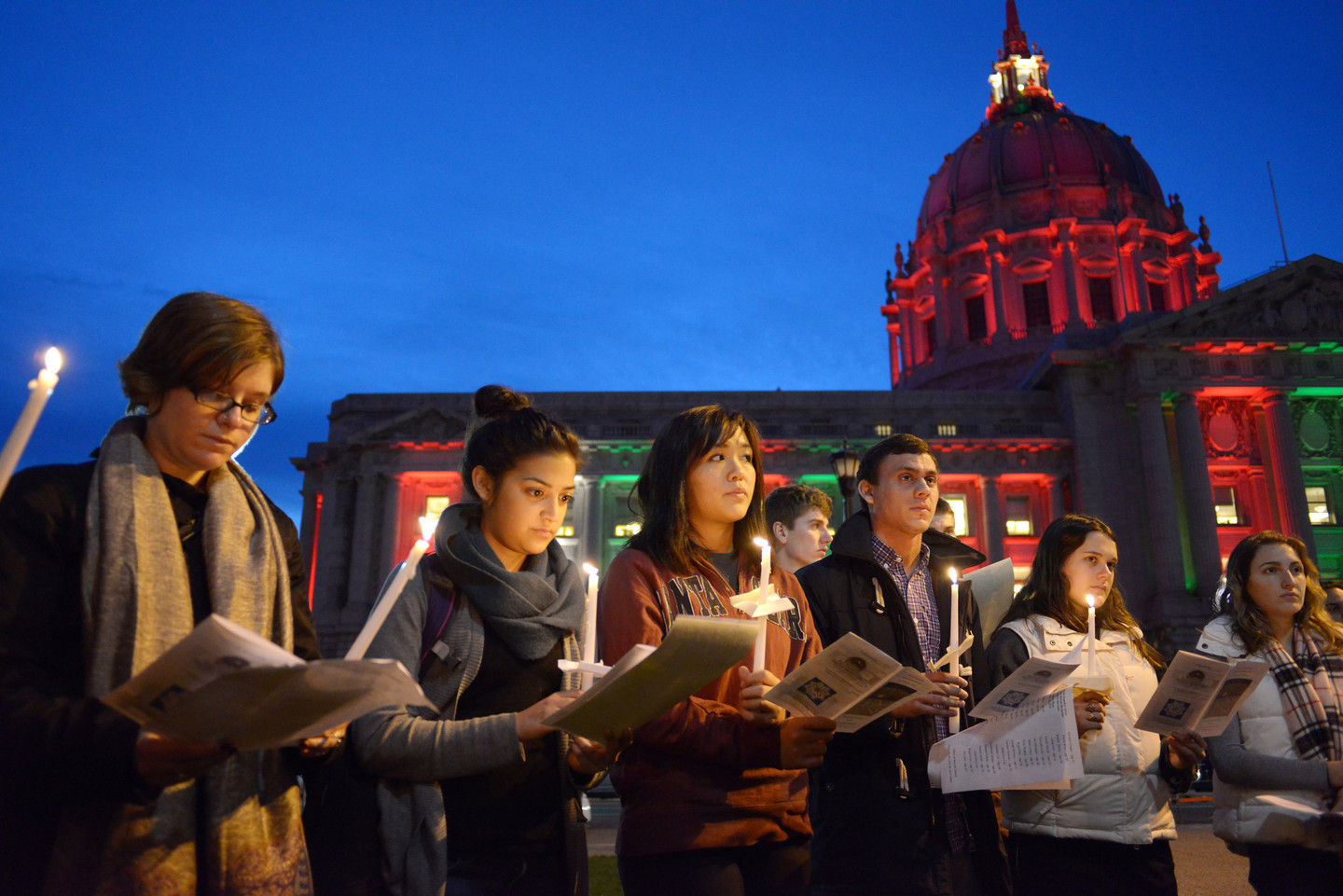 A line of six people, all presenting as white or Asian, hold a long, white, lit candle in one hand and what seems to be a paper program in the other. Some look down at the program, and others look up at a focal point beyond and to the left of the camera. Behind them is the south half of the facade of San Francisco City Hall (facing what would be the plaza), the rotunda lit up in red, and the wall beneath it lighted in alternating columns of red and green. The sky, which takes up most of the top half of the photo, is a deep blue, as after sunset.