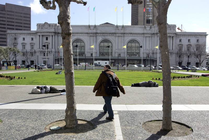 Daytime in San Francisco's Civic Center Plaza, with the monolithic Bill Graham Auditorium (a block-long, maybe two-story building with a cement brick face) as the backdrop. A man strides between the trunks of two trees, his long shadow falling behind him on one of them, toward two figures lying on the sidewalk, at the edge of a green lawn.