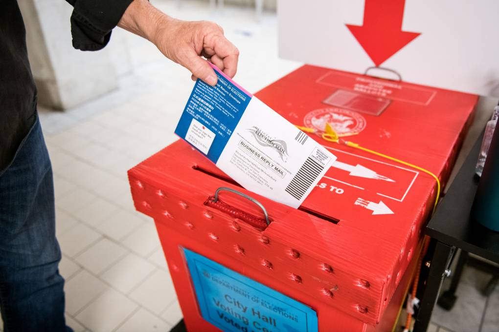 A hand places a vote-by-mail ballot into a bright red ballot box.