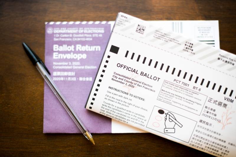 A ballot and its envelope on a dark wooden table, with a pen laid on top of them, photographed from above
