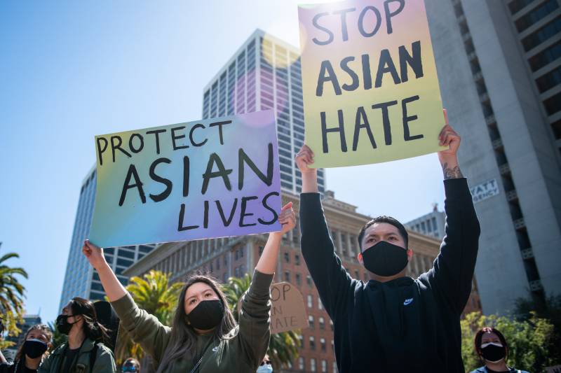 A man and a woman wearing black face masks hold up signs that read "Protect Asian Lives" and "Stop Asian Hate" outside in a crowd.