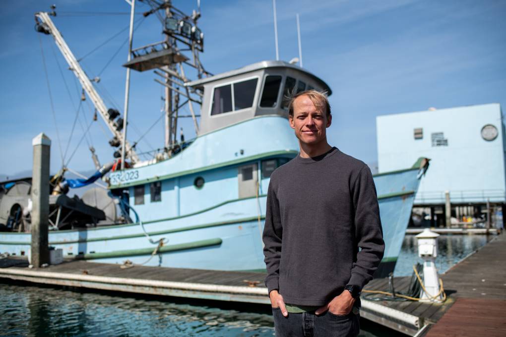 A man wearing a black shirt stands in front of a light blue boat on a dock.