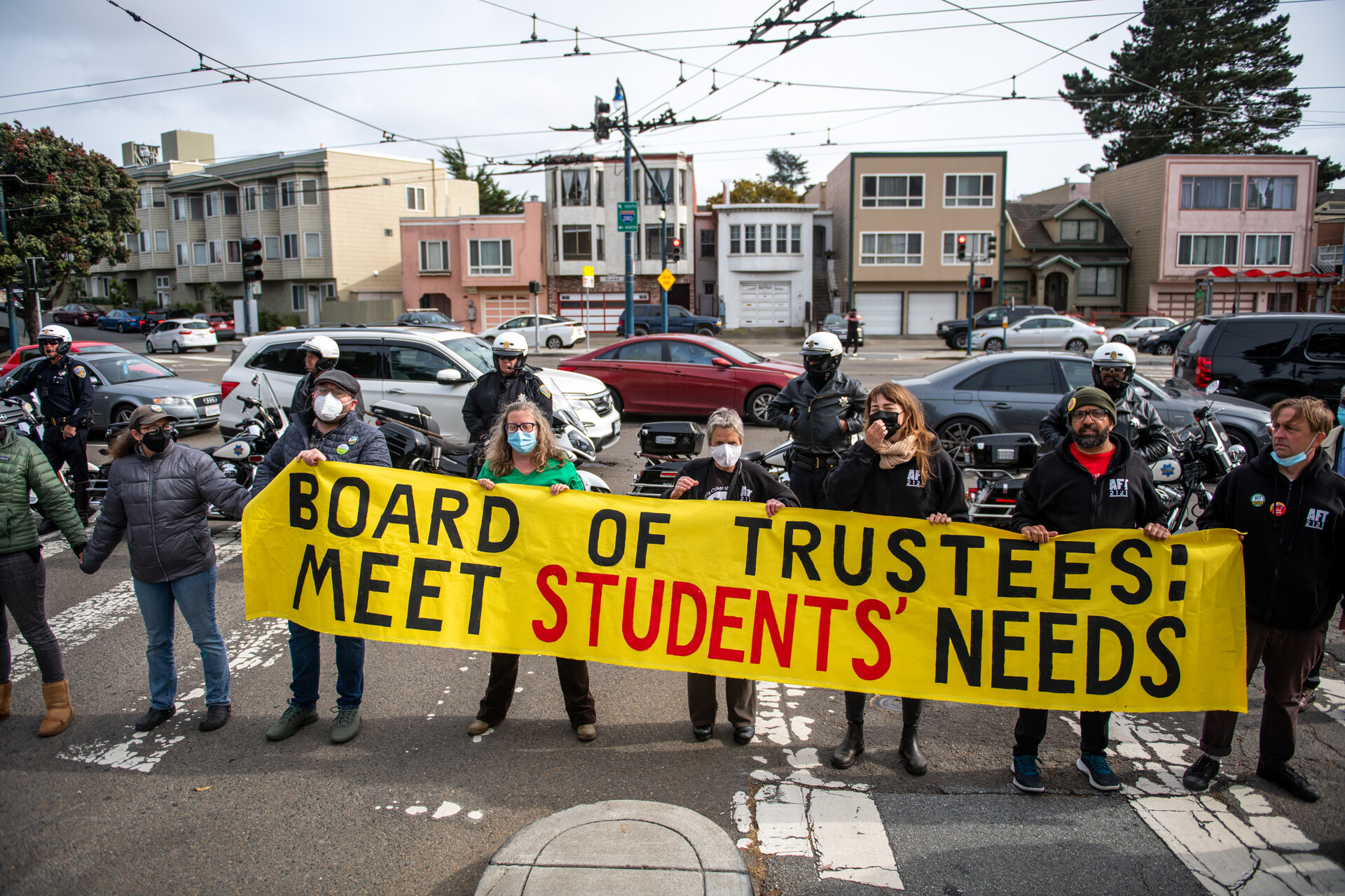 Protesters hold large yellow banner sign reading 'board of trustees meet students needs'