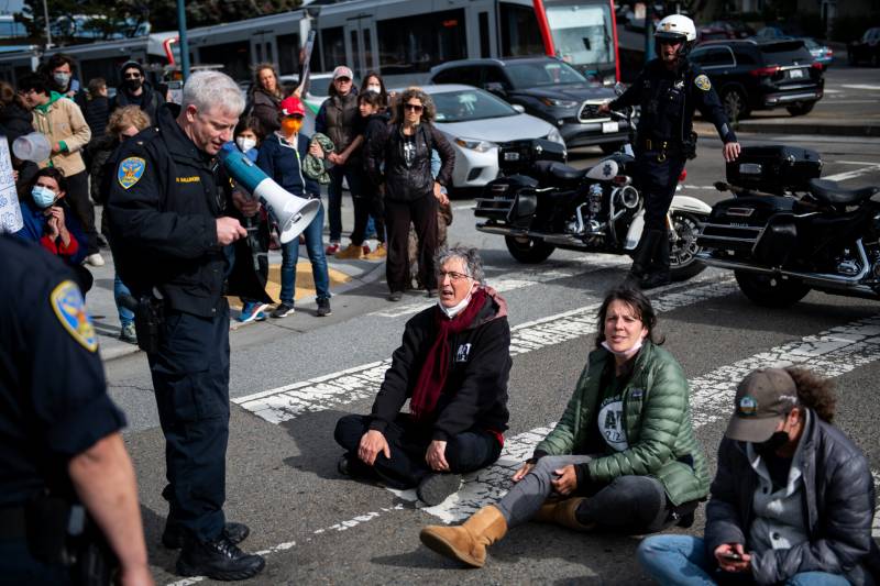 SFPD officer holds megaphone pointing down to three protesters sitting cross legged in the street as police and other protesters look on