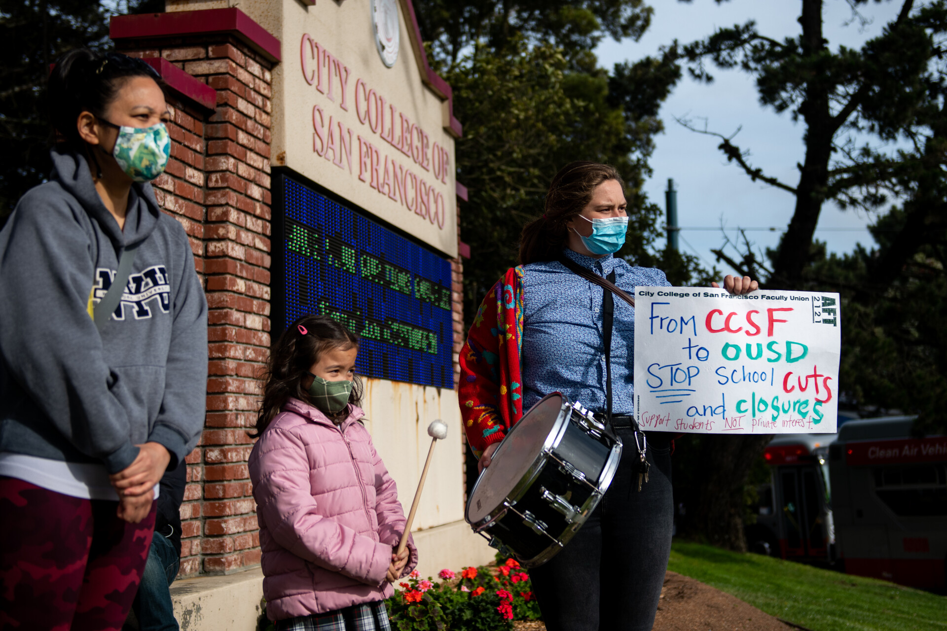 two women and a young girl stand in front of a CCSF sign holding a drum and their own protest sign reading 'from CCSF to OUSD, stop school cuts and closures'