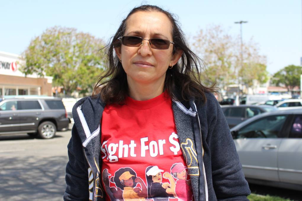 A woman wearing sunglasses and a red shirt that says "Fight for $15" stands in a shopping center parking lot.