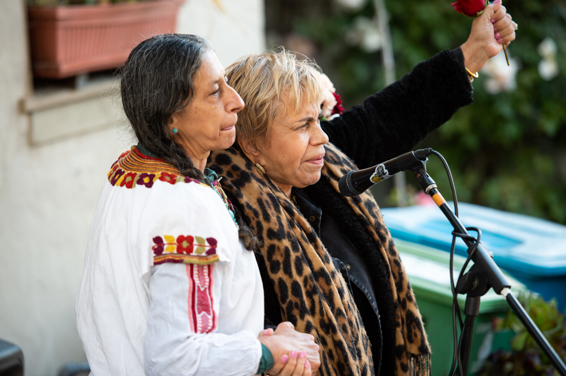 Two women stand together in front of a microphone. One woman wearing a leopard print jacket has her fist raised.