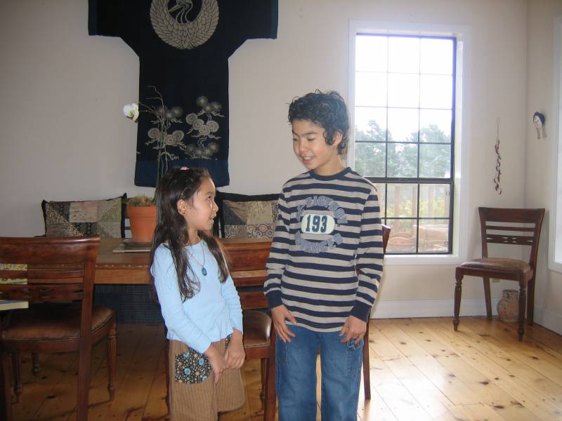 A young girl and boy stand in a living room, with a Japanese garment hanging on the wall behind them.