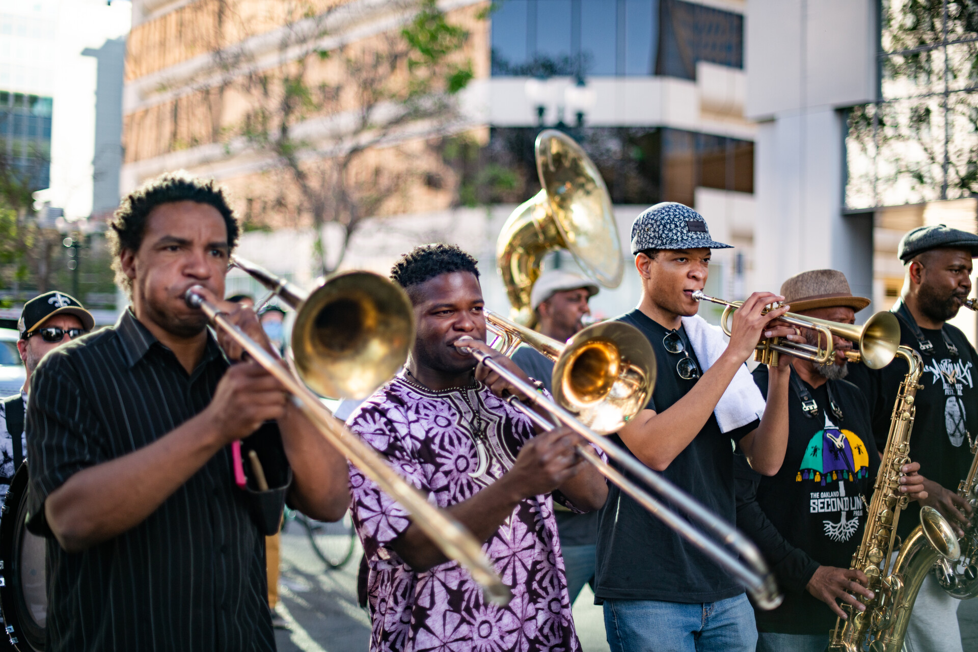 Line of Black men playing trombones, trumpet and tuba walk down a street on a sunlit evening