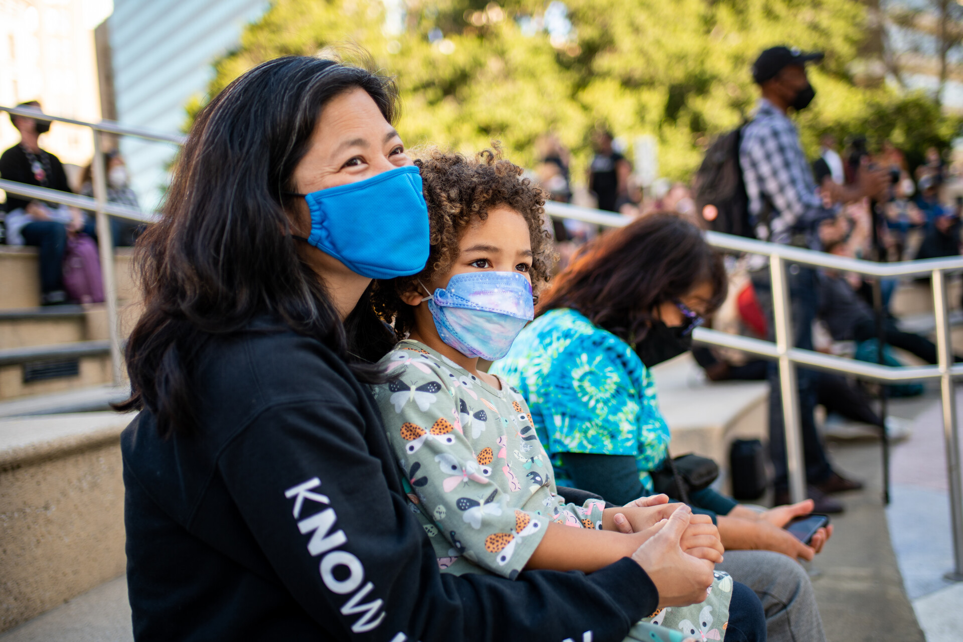 masked woman with smiling eyes holds her young daughter also wearing a mask outside, with other attendees in the background