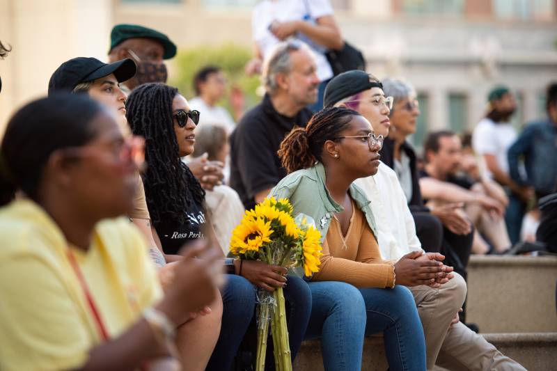 A group of mostly Black people sit outside on steps with solemn expressions, listening to speakers - one of them holds sunflowers