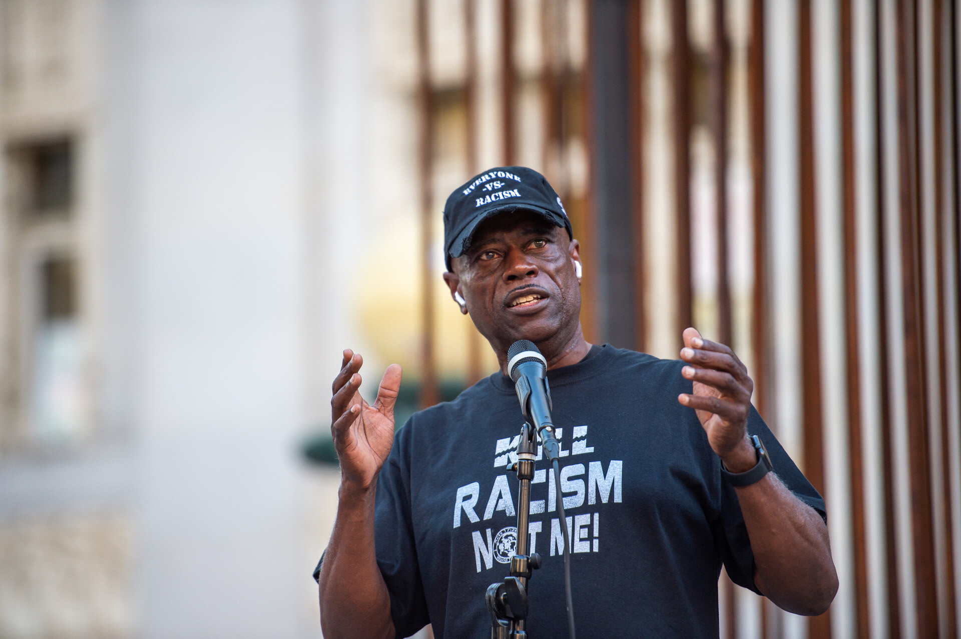 An older Black man speaks at a microphone with a shirt reading 'kill racism not me'