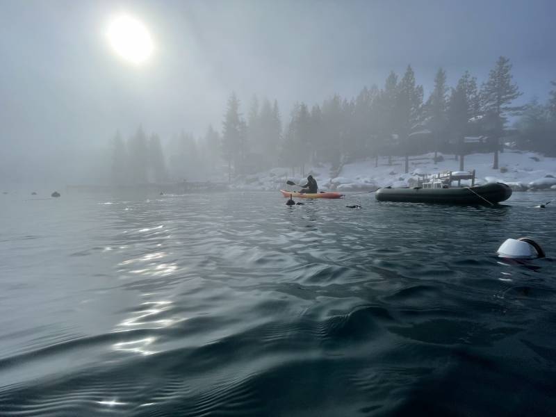 A man in a yellow kayak tows a large rubber raft filled with trash on the rippling black surface of Lake Tahoe in winter. Pine trees dot a snow-covered hill next to the lake. A pale sun shines through fog and makes a white trail along the water. 