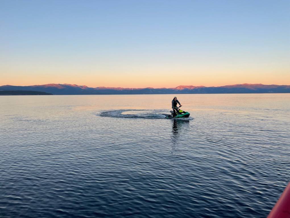 A man on a jet ski swirls his craft around on the blue-black surface of Lake Tahoe at sunset. In the distance, sun lights the tops of dark blue mountains with pink and gold. A band of orange and yellow stretches across the horizon, under a pale blue sky.