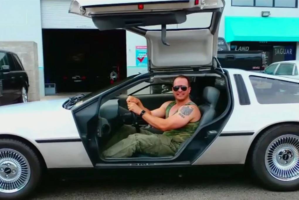 man wearing camo shirt with bare arms and visible Nazi-esque eagle tattoo sits at wheel of Delorean car with door open