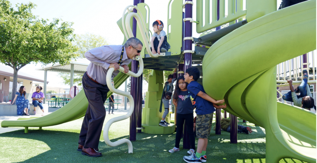 A man wearing glasses, dress pants and a dress shirt leans with both hands on a green playground apparatus talking to two children.