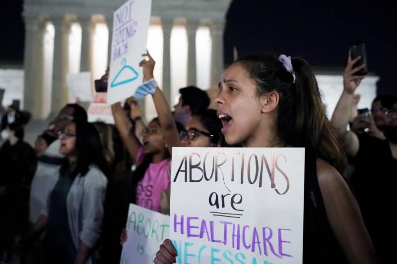 A group of protesters outside the Supreme Court, with mouths open in speech or chanting. The woman closest to the camera is holding a sign saying "Abortions are healthcare."