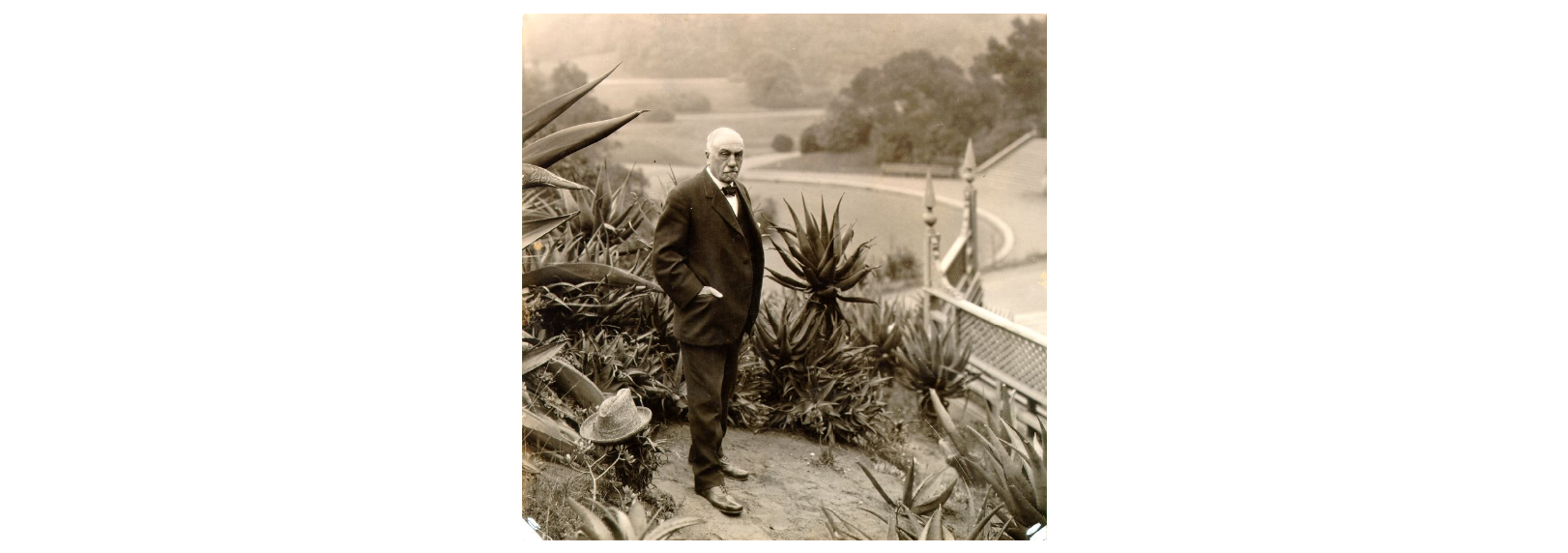 Sepia toned photo of a tall man in a black suit with large eyebrows. He stands surrounded by palm trees with more greenery behind him.