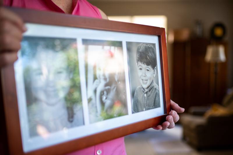 A rectangular wooden frame holds three images of children. Two are blurred and the third shows Kory as a young boy in a denim shirt with a mop of dark hair, black eyes, and a happy smile.