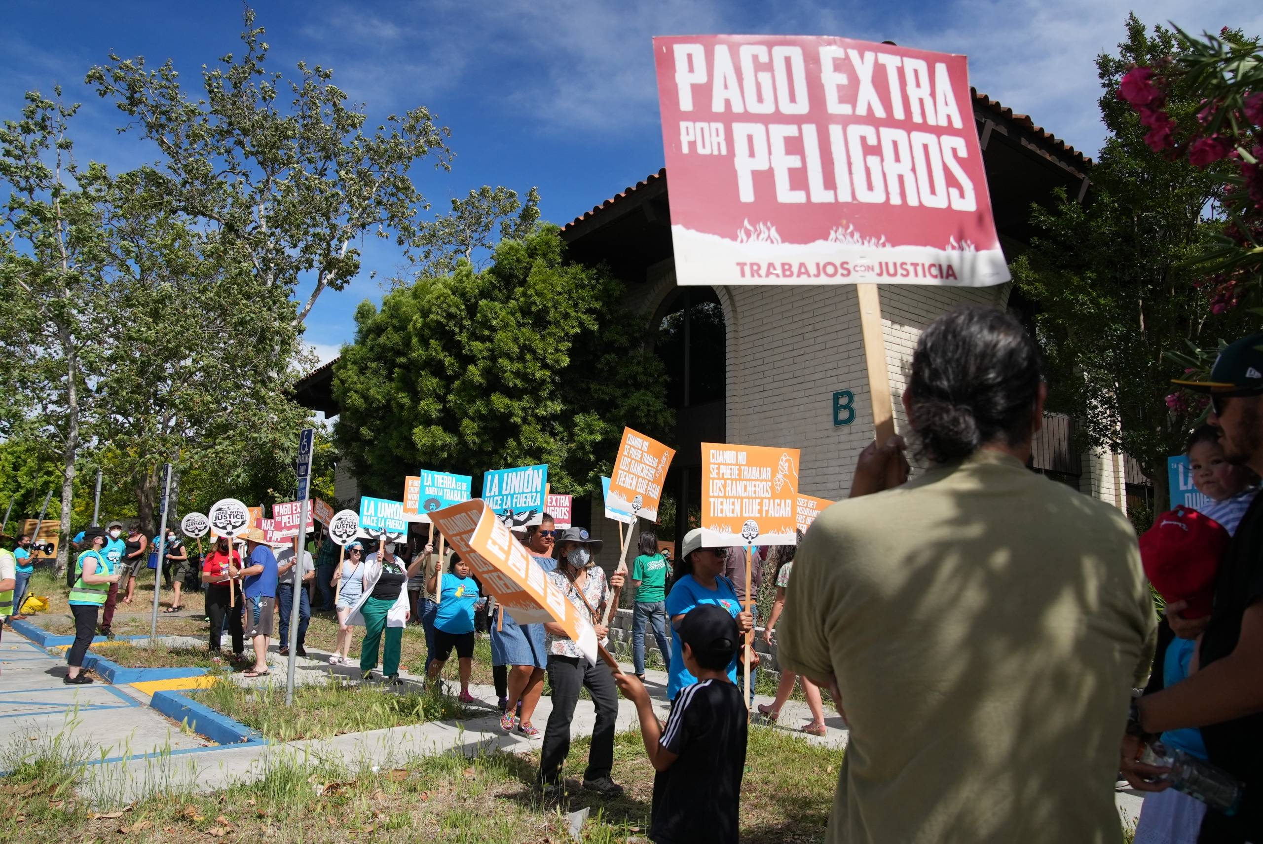 A group of people protest outside an office building. Many are holding signs, one says in Spanish, "Pago extra por peligros" or "hazard pay" in English.