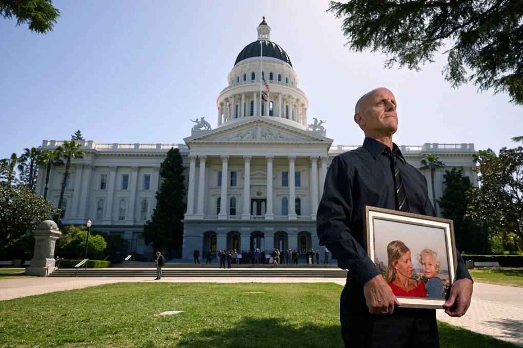 Douglas Forbes stands in front of the California Capitol holding a painting of his late wife and his daughter
