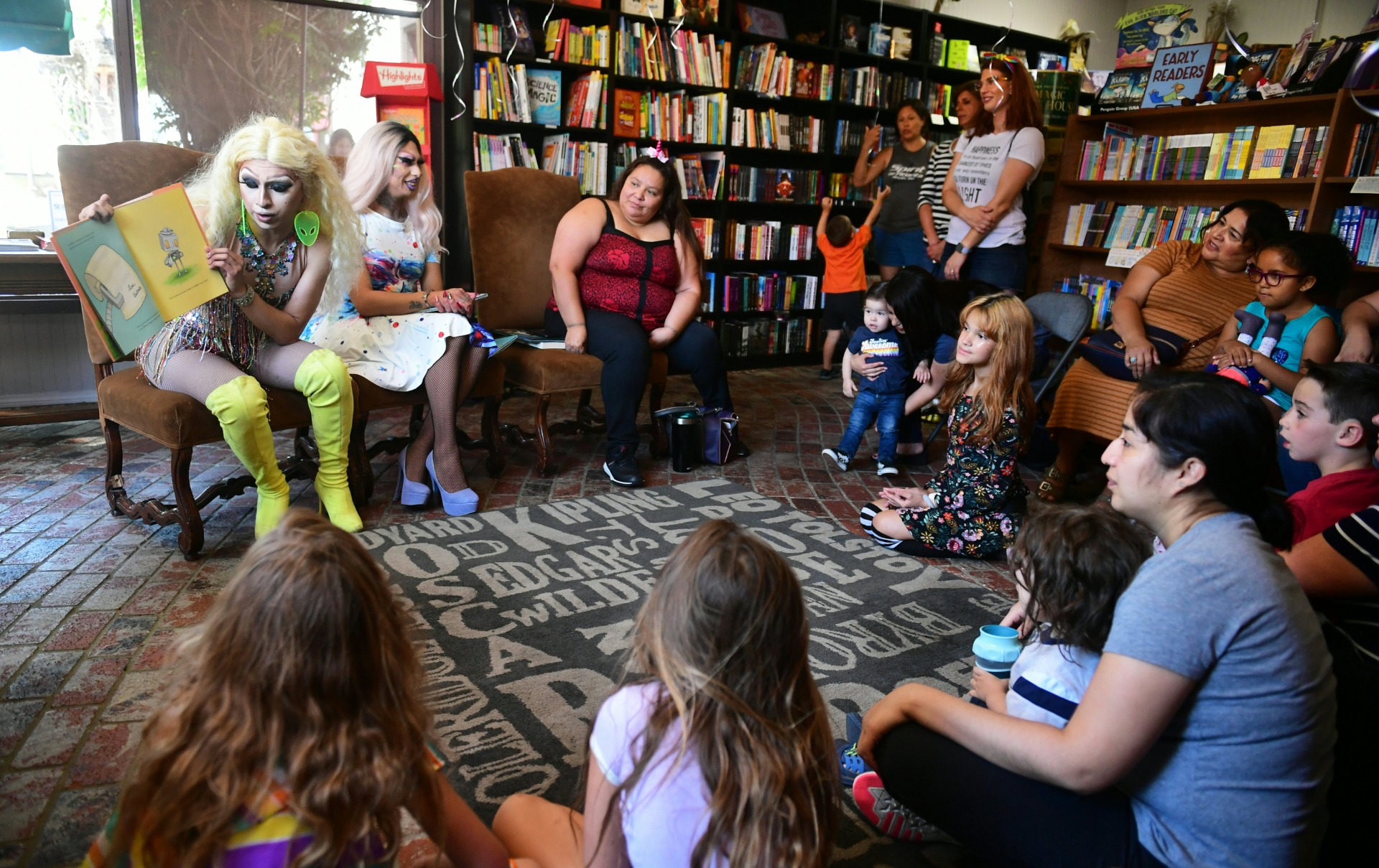 A drag queen, left, holds open a storybook and reads for children sitting in a circle around them inside a library.