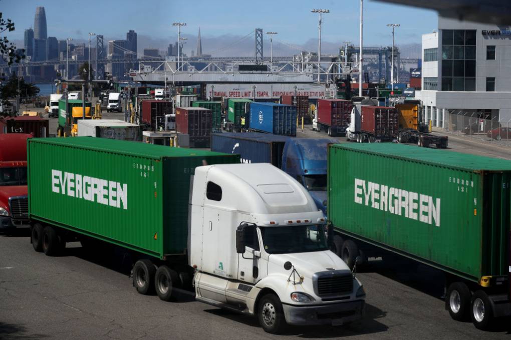Two bright green containers on flatbed trucks (the white cab of one truck is visible), with lettering on the side that says "Evergreen" in white, sit in a shipping yard. Around them are many other flatbeds, carrying mostly blue or red containers. In the distance, on the left, you can see a sliver of the San Francisco Bay; the Bay Bridge crosses the entire frame, and beyond that is the SF skyline, including the Salesforce and Transamerica towers.