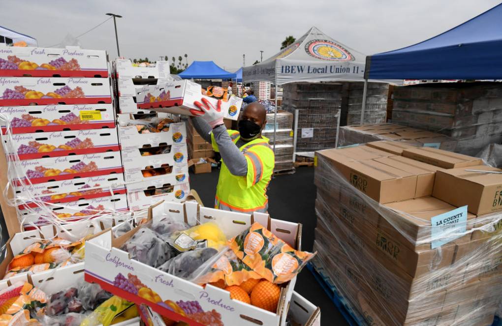 An outdoor scene, likely in a parking lot, on an overcast day. A bald middle-aged Black man wearing a black face mask, white gloves with orange rubber palms, and a neon yellow vest over a gray long-sleeved shirt hoists a white, open-top, cardboard box above his head, taking it down from a tall pile of similar boxes. He is surrounded by towers of these plastic-wrapped boxes; a box in the foreground shows that they're full of fruit: oranges, and what looks like lemons and plums. Behind him, beneath at least four blue tents/shade structures, are more towers of plastic-wrapped brown cardboard boxes -- we can't see inside them. A gray tent says "IBEW Local Union 11," below a circular crest of concentric circles of black, red, blue and then yellow in the middle. Way in the distance are the tippy-tops of a row of coconut palms, and a utility pole.